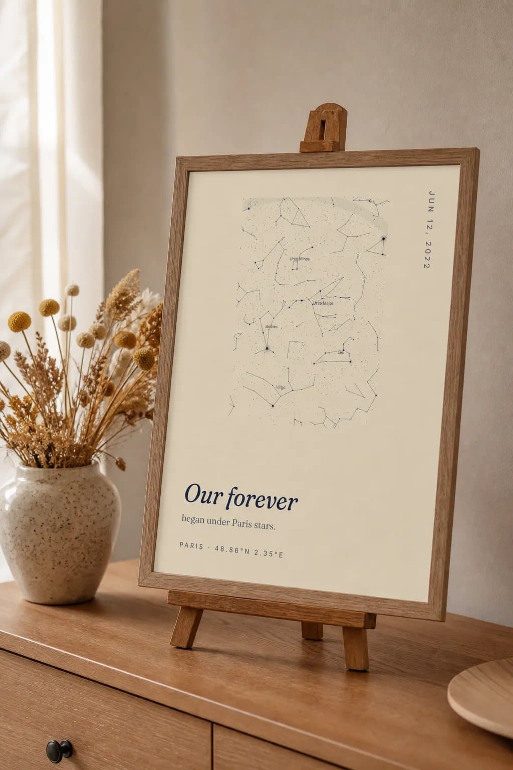 A framed cream star map titled Our forever standing on a small wooden easel beside dried flowers.